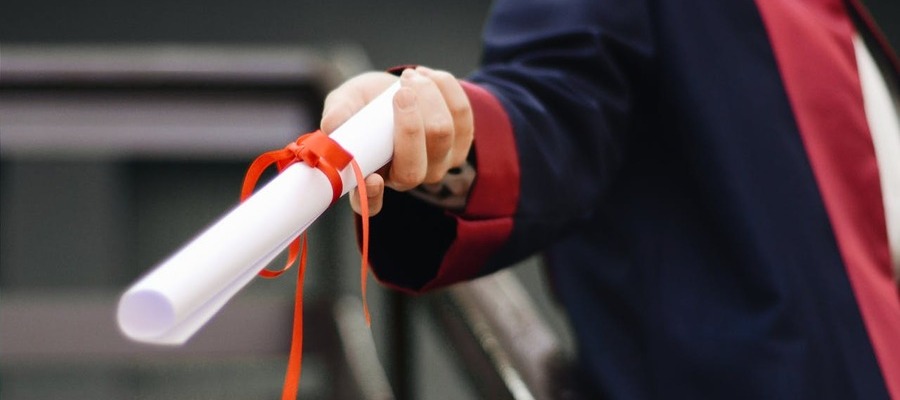 A graduate wearing a gown and holding a diploma tied with a red ribbon.